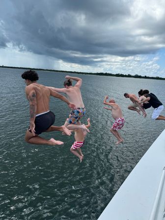 Six people jumping off a boat into a bay, mid-air over rippling water in colorful swim trunks with dramatic dark storm clouds overhead