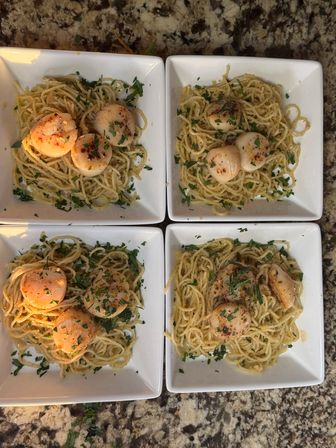Four white square plates of angel-hair pasta topped with seared scallops and chopped parsley, plated as a seafood dinner on a speckled granite countertop.