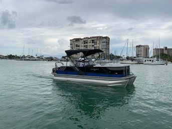 Blue and silver pontoon boat cruising in calm marina waters with sailboats and waterfront condominium buildings under an overcast sky