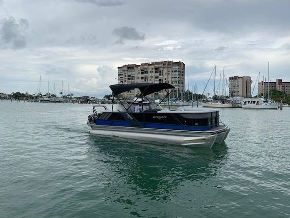 Blue-black pontoon boat cruising calm teal harbor waters near a marina with sailboats and waterfront condos under an overcast sky.