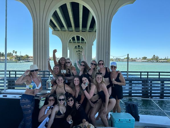 Cheerful group of women in swimsuits posing on a dock under a seaside concrete bridge arch, turquoise water and palm trees in the background on a sunny day.