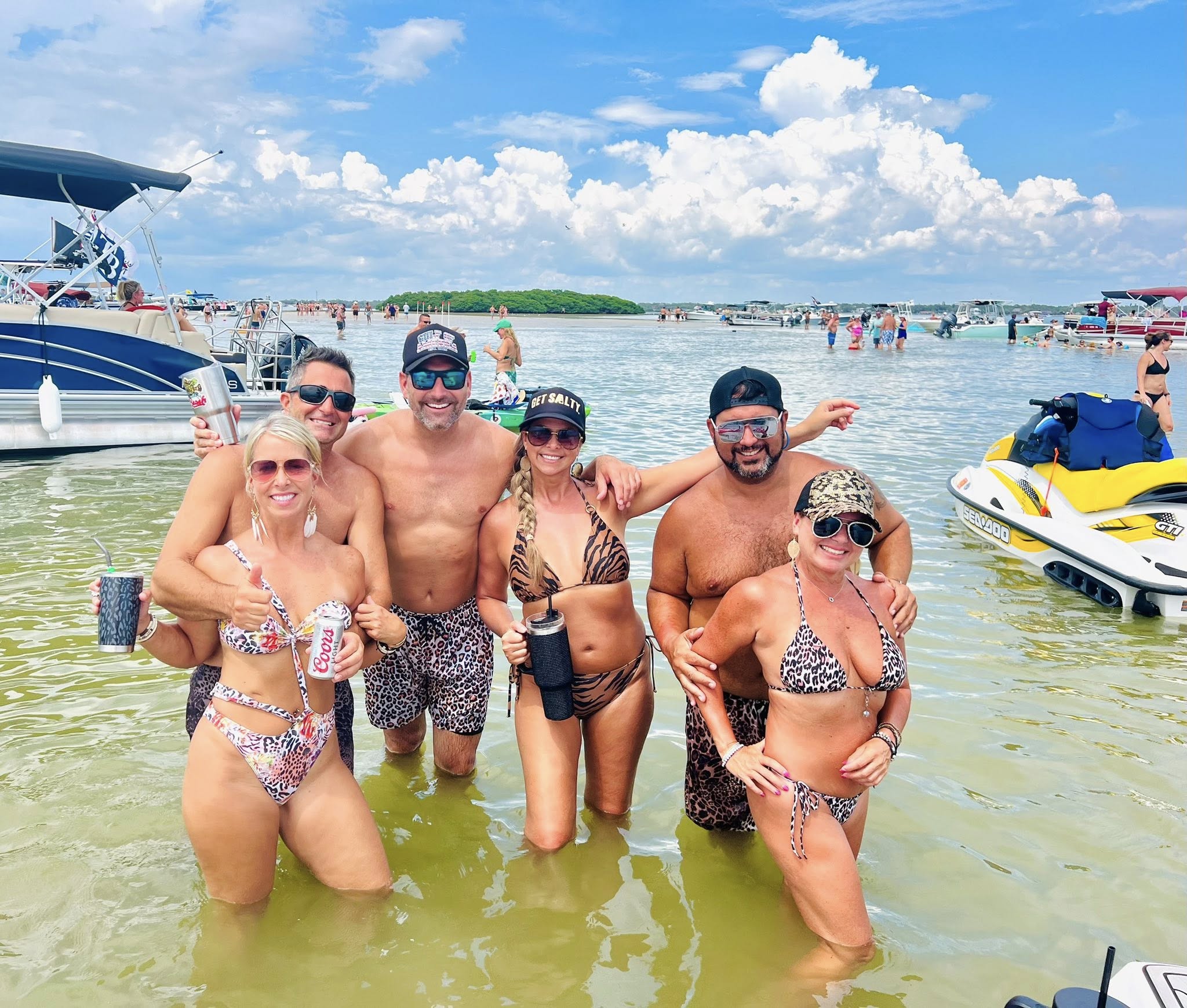 Six friends in swimsuits posing waist-deep at a busy sandbar with anchored boats and a yellow jet ski under a sunny blue sky.