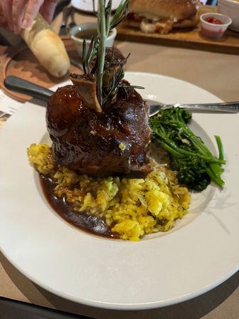 Hearty braised beef shank with exposed marrow bone and rosemary sprig, sitting on yellow risotto with broccolini and a glossy brown sauce on a white dinner plate.