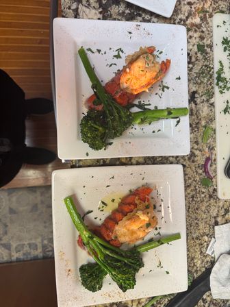 Two chef-plated grilled lobster tails with broccolini on white square plates, garnished with chopped parsley and a light drizzle on a granite countertop — elegant seafood presentation.