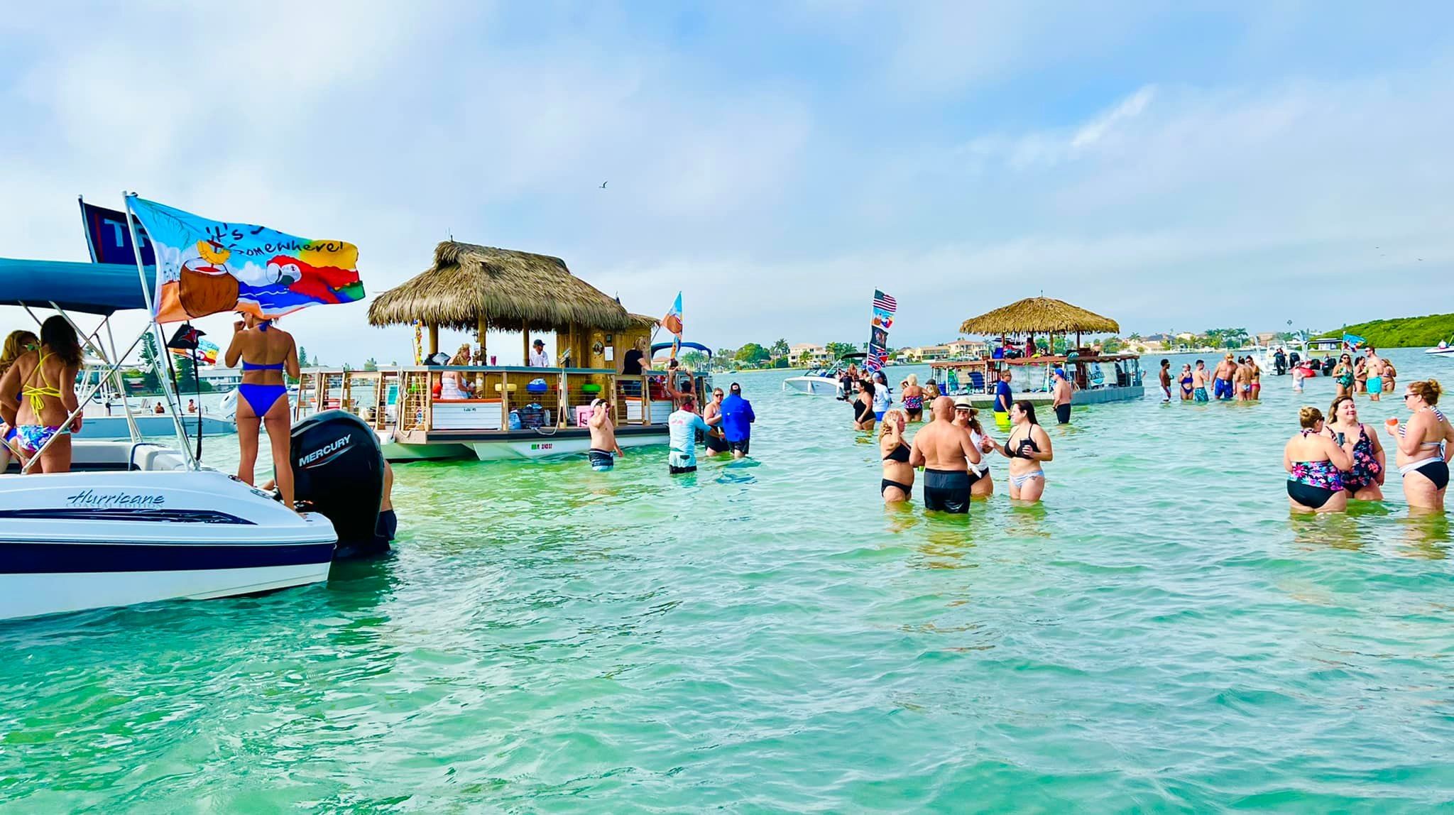 Crowded sandbar boat party in shallow turquoise water with people wading and socializing around boats and thatched-roof floating tiki bars under a sunny blue sky