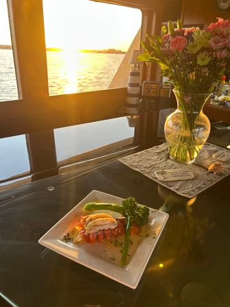 Plated lobster tail with broccolini on a glossy table beside a vase of flowers in a yacht cabin, golden sunset reflecting across calm water through the window — waterfront sunset dinner scene.