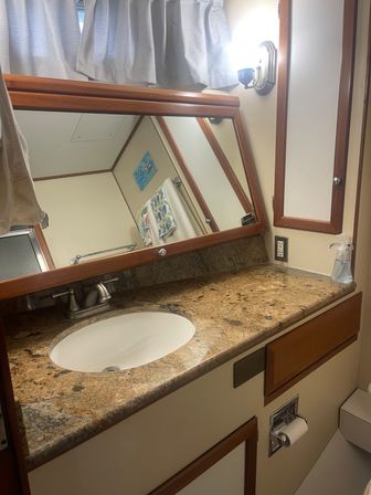 Cozy compact marine-style bathroom vanity with brown granite countertop, oval undermount sink, brushed-nickel faucet, wood-trimmed mirror and cabinets, wall sconce, towel reflected in mirror and soap dispenser.