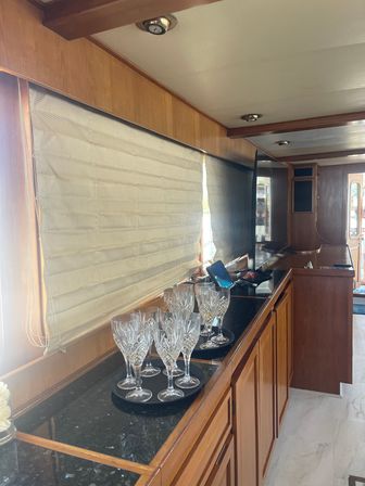 Yacht cabin interior with warm wood paneling and black granite counter, sparkling crystal wine glasses on trays, beige blinds partially drawn and a wall-mounted TV nearby
