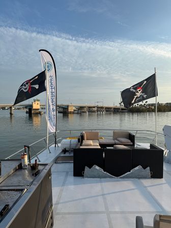 Boat deck lounge with wicker sofas and two Jolly Roger pirate flags flying near a coastal drawbridge over calm water under a pastel sky