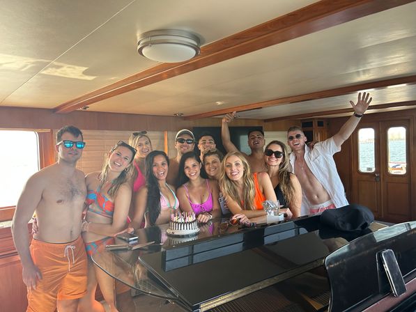 Friends in colorful swimwear celebrating a birthday around a cake on a black piano inside a sunny yacht cabin, ocean visible through the windows