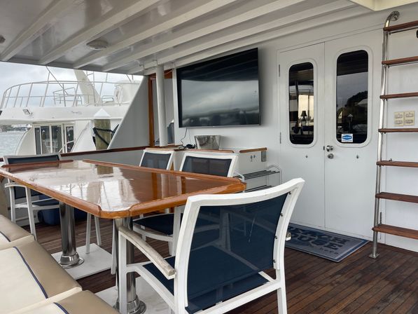 Aft deck dining area on a luxury motor yacht with glossy teak table, white-frame navy mesh chairs, wall-mounted TV, ladder to flybridge and marina view