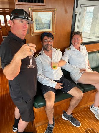 Three people relaxing aboard a wood-paneled yacht cabin — two crew in white captain shirts with epaulettes and a guest in black, toasting with drinks on a green bench beside nautical charts and an ocean view.