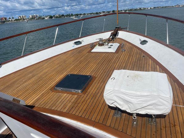 Sunlit teak-deck bow of a yacht with covered gear and cleats, calm harbor waters and coastal skyline under a partly cloudy sky.