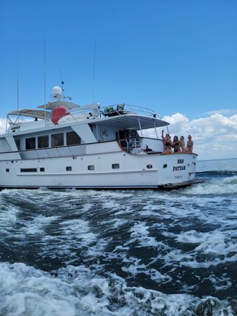 White motor yacht cruising on choppy blue sea under a clear sky, four people in swimsuits waving from the stern with foamy wake trailing behind.