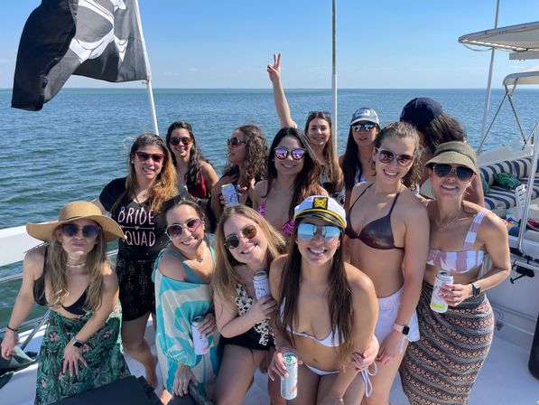 Group of women friends on a summer boat in the ocean celebrating a bachelorette party — swimsuits, sunglasses, captain’s hat, canned drinks and a pirate flag.