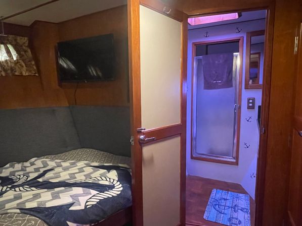 Cozy wooden boat cabin with navy nautical bedding, wall-mounted TV, and open door to a compact bathroom with frosted shower door, towel, and anchor-themed decor.