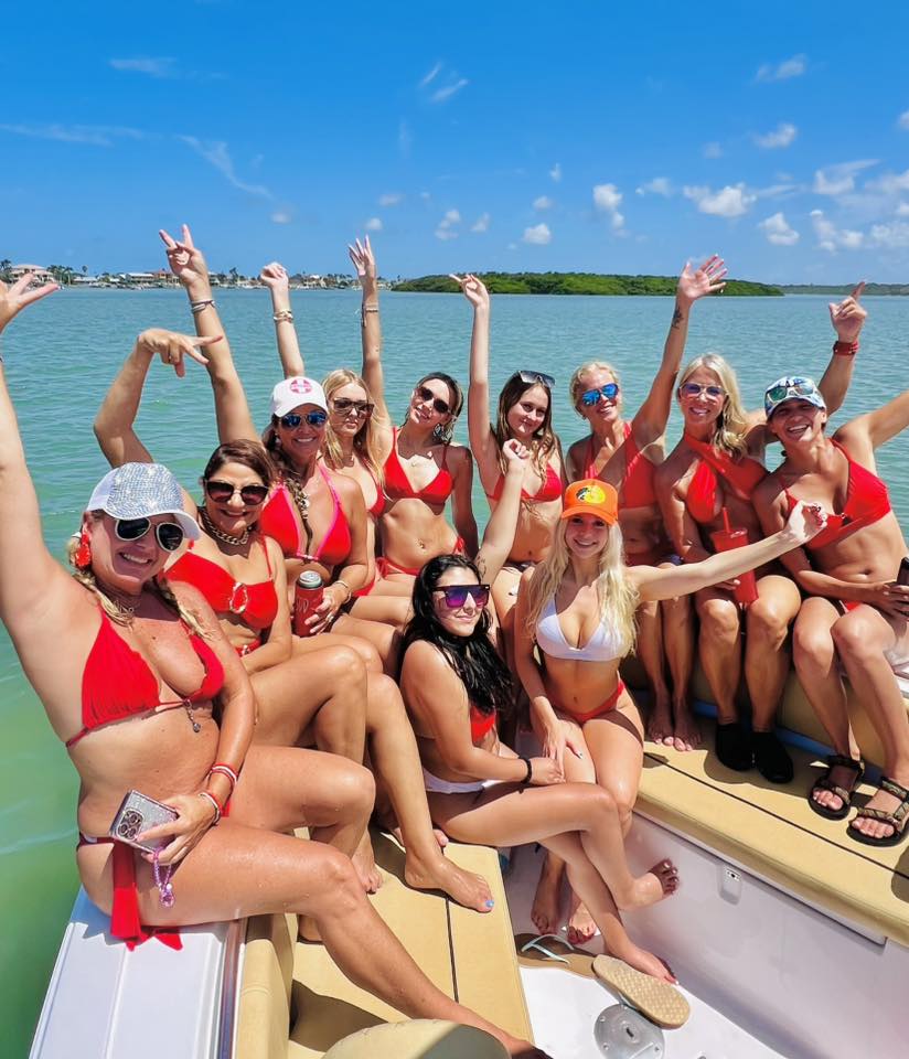 Energetic boat party with a group of women in red bikinis cheering and posing on a sunny day over turquoise coastal waters with a blue-sky island backdrop.
