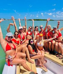 Energetic boat party with a group of women in red bikinis cheering and posing on a sunny day over turquoise coastal waters with a blue-sky island backdrop.