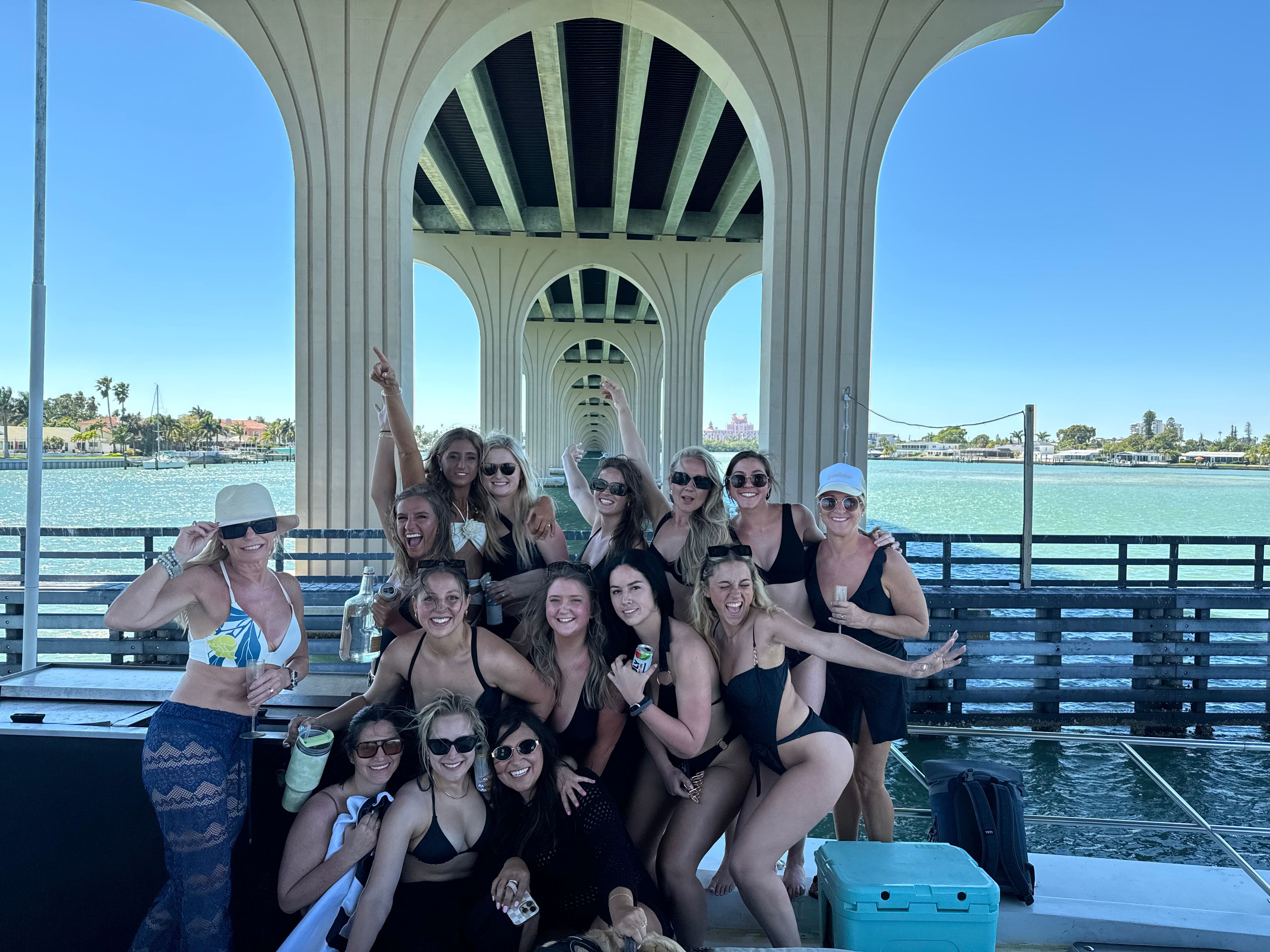 Group of women in swimsuits laughing and posing under a concrete arched waterfront bridge, turquoise bay and clear blue sky in the background with a cooler and drinks nearby — sunny pier/boat gathering.