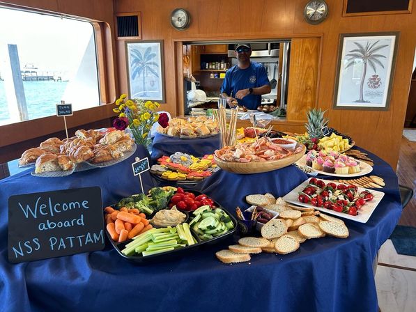 Colorful buffet spread aboard a yacht with seafood, pastries, veggie platters and desserts, sea view through a window, welcome aboard chalkboard and crew member in the galley