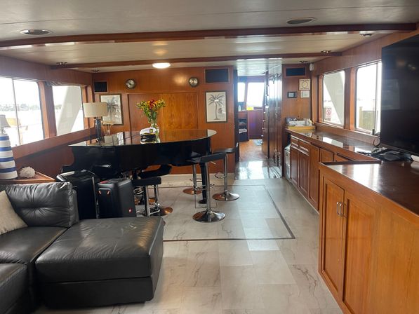 Sunlit yacht salon interior with a black baby grand piano doubling as a bar with stools, leather sectional, wood-paneled walls and cabinetry, large windows overlooking a marina, and fresh flowers on the piano.