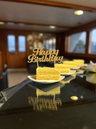 Slice of yellow layered cake on a white plate with a glittery "Happy Birthday" topper reflected on a glossy black surface, arranged in a row inside a warm wooden yacht cabin — elegant birthday dessert display.