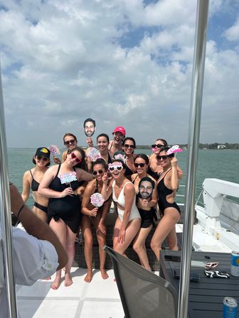 Bachelorette party group of women in swimsuits posing on a yacht over calm coastal waters, holding playful signs and oversized face cutouts under a partly cloudy sky.