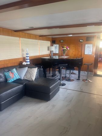 Cozy yacht lounge with black leather sectional, glossy black grand piano doubling as a bar with chrome stools, striped nautical lamp, vase of fresh flowers and palm-print art on warm wood-paneled walls.