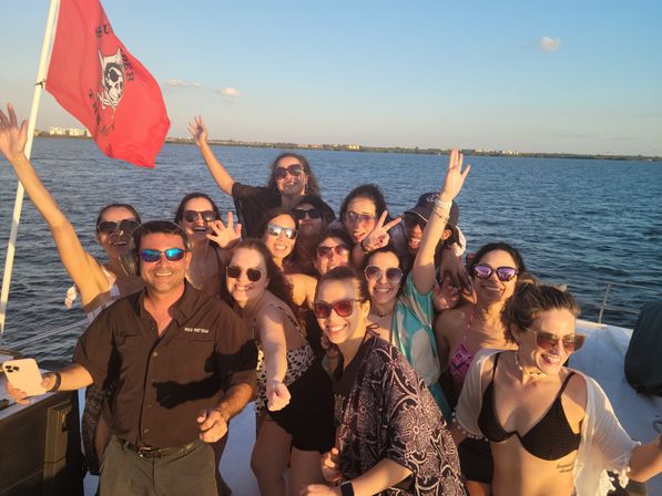 Energetic boat party group posing and smiling at golden-hour on a coastal sunset cruise, people in sunglasses and swimwear with a red pirate flag and waterfront skyline in the background.