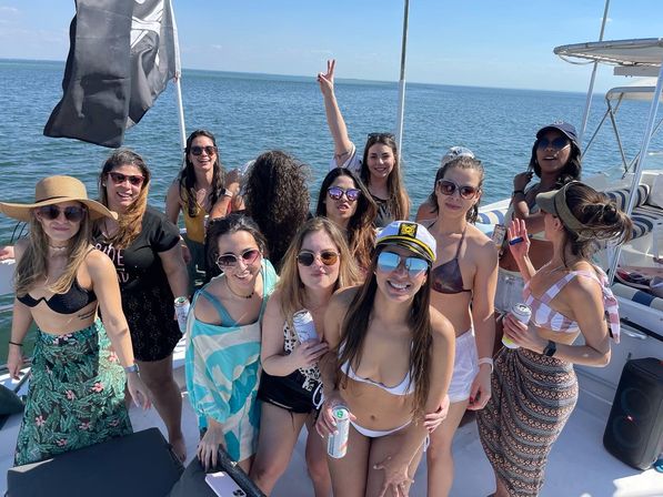 Smiling group of women in swimsuits on a sunlit boat party over calm blue water, holding drinks and one wearing a captain's hat.