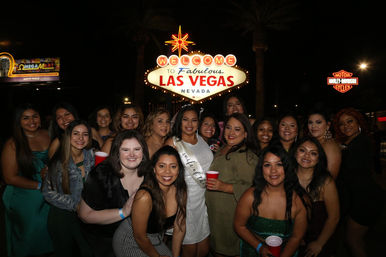 Smiling group of women posing at night in front of the illuminated "Welcome to Fabulous Las Vegas, Nevada" sign, many holding red cups and celebrating.