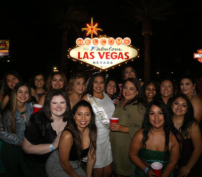 Smiling group of women posing at night in front of the illuminated "Welcome to Fabulous Las Vegas, Nevada" sign, many holding red cups and celebrating.