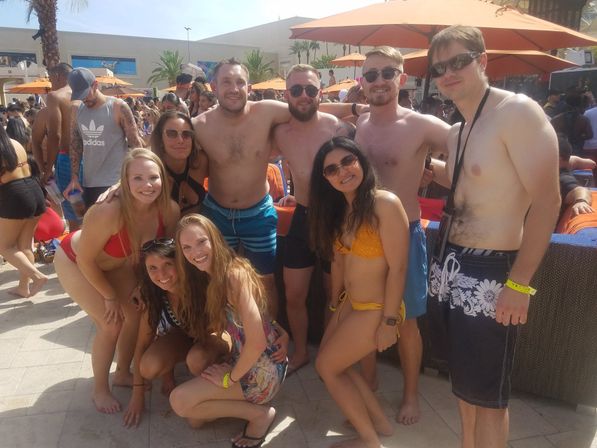 Group of friends posing at a sunny outdoor pool party in swimwear, orange umbrellas and palm trees lining a lively resort pool scene.