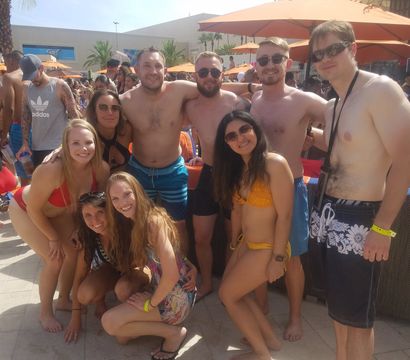 Group of friends posing at a sunny outdoor pool party in swimwear, orange umbrellas and palm trees lining a lively resort pool scene.