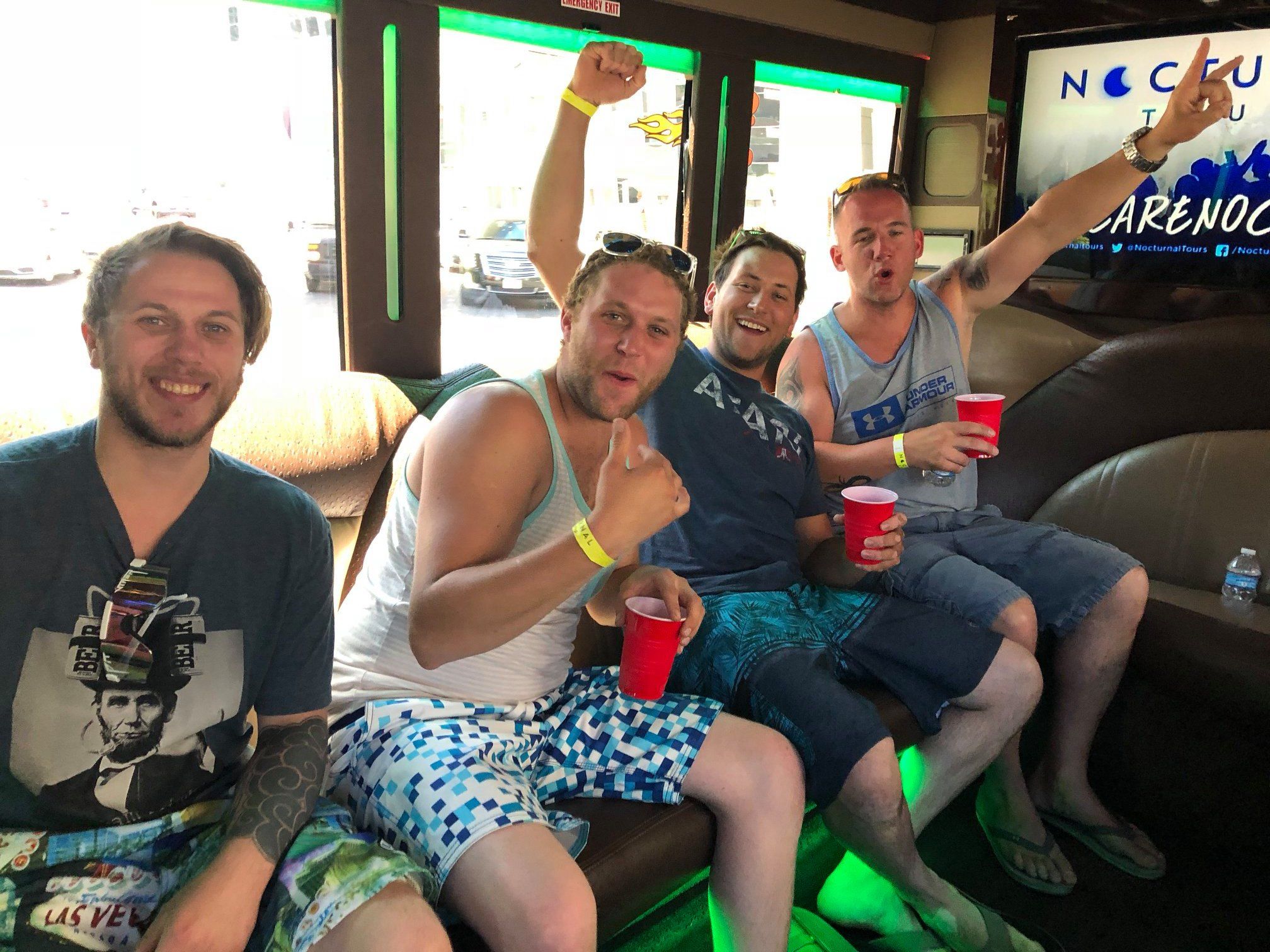 Four friends smiling and cheering on a neon-lit party bus in Las Vegas, wearing summer clothes and flip-flops and holding red cups for a casual daytime celebration.