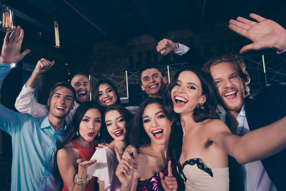 Group selfie of young adults celebrating at an indoor party or nightclub, smiling, cheering, and posing under warm decorative lights