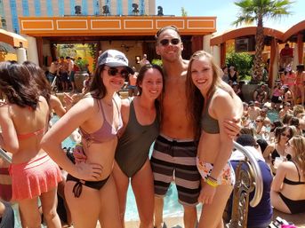 Four young adults smiling in swimsuits at a crowded outdoor resort pool party with cabanas, palm trees, and sunny weather.