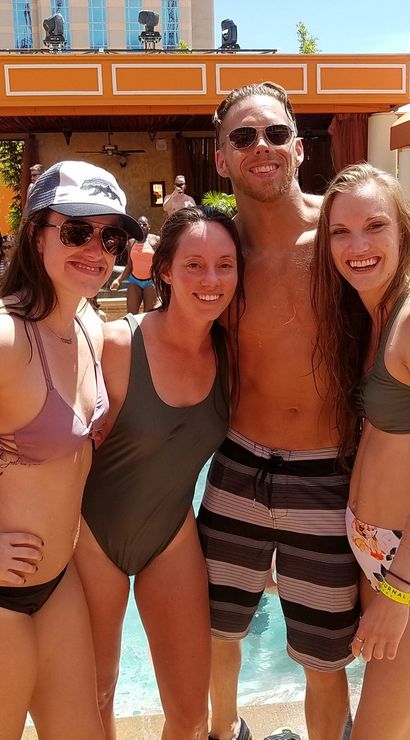 Four young adults smiling in swimsuits at a crowded outdoor resort pool party with cabanas, palm trees, and sunny weather.