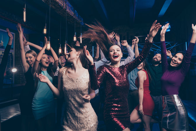 Energetic nightclub dance party with friends in sparkly dresses and casual outfits dancing and cheering under hanging Edison bulbs on a lively night out