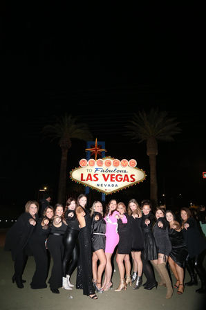 Nighttime group of women posing and pointing at the camera in front of the illuminated "Welcome to Fabulous Las Vegas Nevada" sign with palm trees, lively Las Vegas celebration