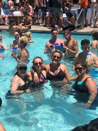 Crowded outdoor pool party at a sunny resort-style pool, friends in swimwear smiling and sipping colorful drinks while socializing in waist-deep water