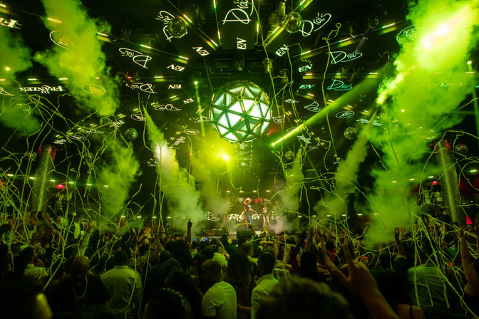 Packed nightclub dance floor with DJ on stage, vivid green lasers and smoke, geometric LED disco ball, and streamers raining down on a cheering crowd.