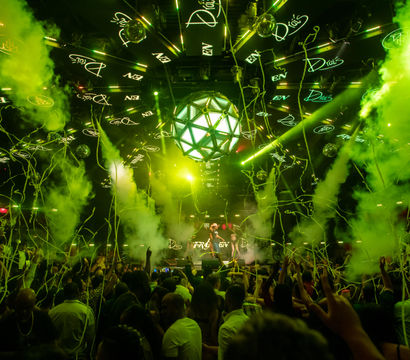 Packed nightclub dance floor with DJ on stage, vivid green lasers and smoke, geometric LED disco ball, and streamers raining down on a cheering crowd.