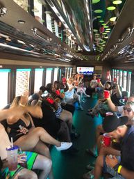 Neon-lit party bus interior with a large group of people seated along both sides, smiling and raising red cups in a lively group celebration.