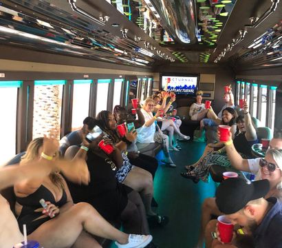 Neon-lit party bus interior with a large group of people seated along both sides, smiling and raising red cups in a lively group celebration.