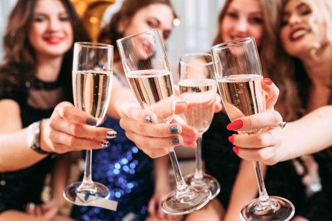 Close-up of champagne flutes clinking in a festive celebration toast by friends wearing sparkly party outfits and colorful nail polish.