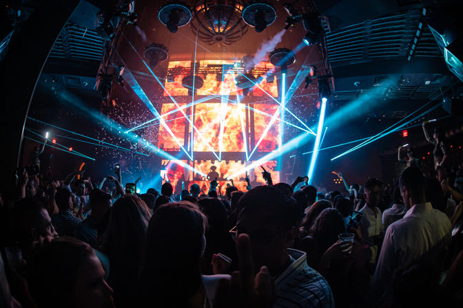 Packed urban nightclub dance floor with a DJ silhouetted against a fiery LED backdrop, blue laser beams and confetti lighting up the lively nightlife crowd