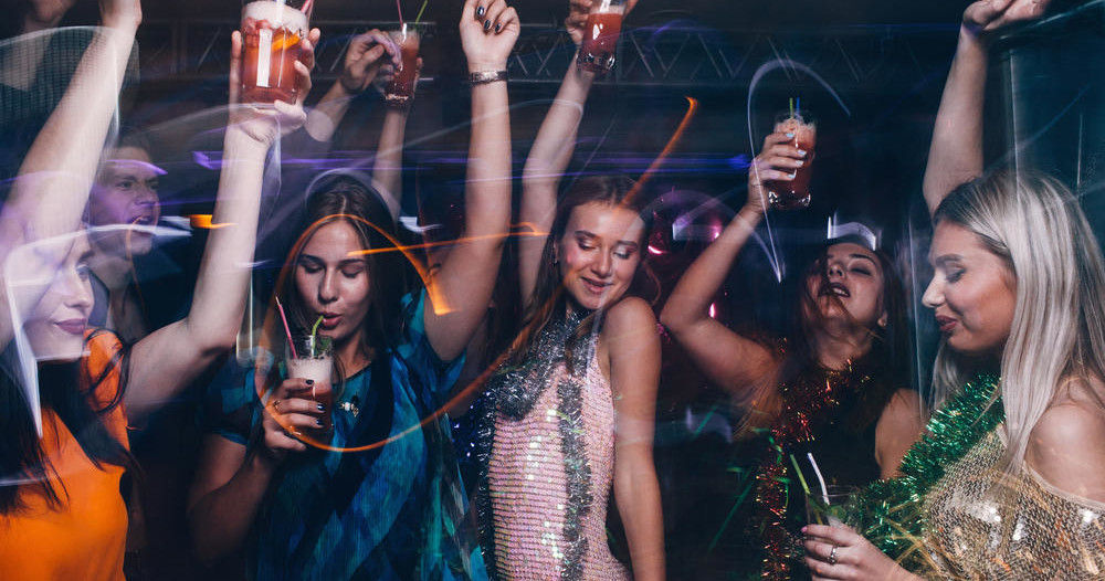 Group of women dancing and raising cocktails on a nightclub dance floor with sequined outfits and colorful party lights