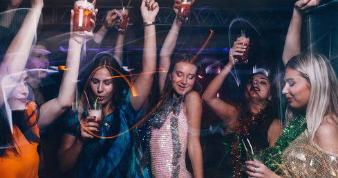 Group of women dancing and raising cocktails on a nightclub dance floor with sequined outfits and colorful party lights