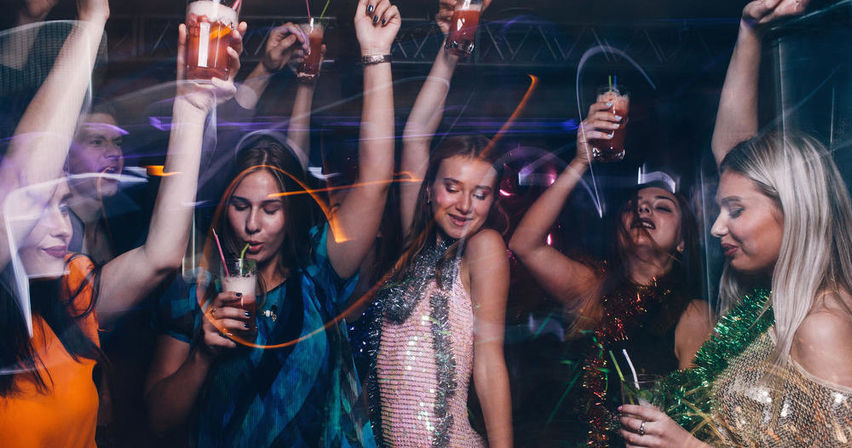 Group of women dancing and raising cocktails on a nightclub dance floor with sequined outfits and colorful party lights